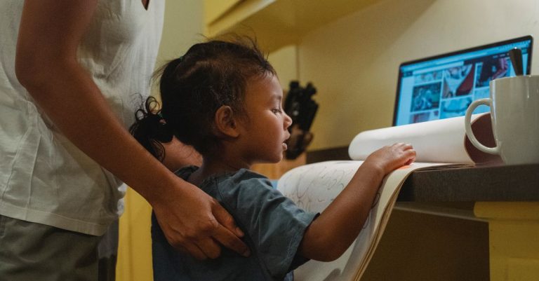 Side view of attentive little ethnic girl showing album with drawings to crop faceless young mother working remotely in laptop at home